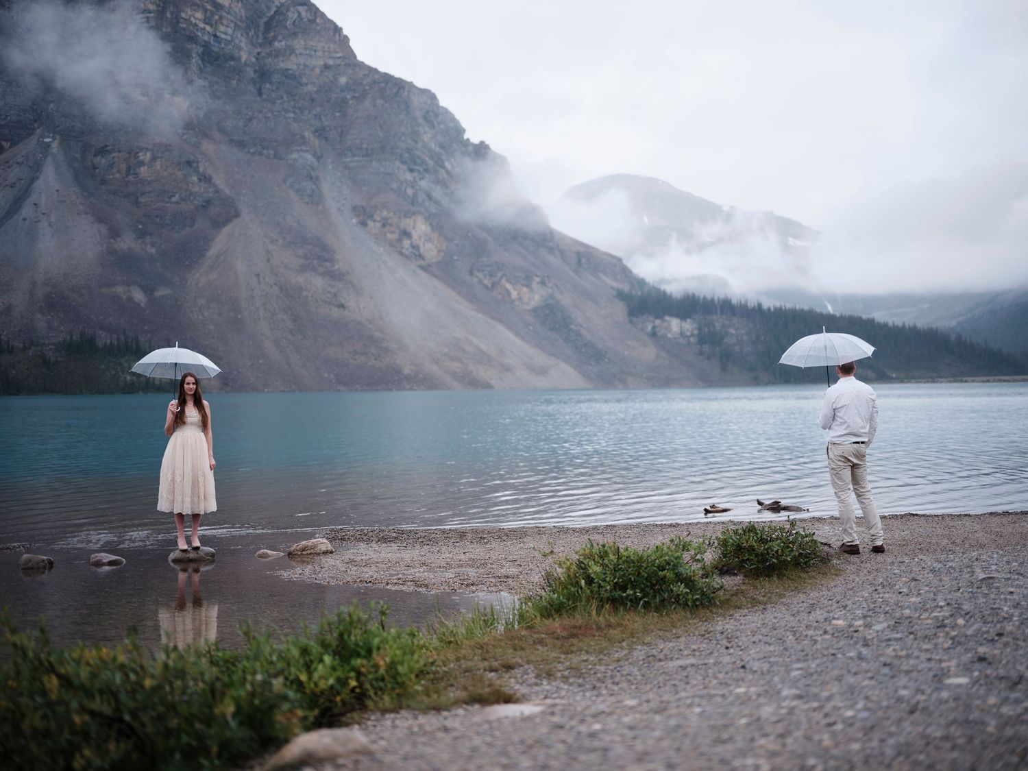 Couple with white umbrellas standing apart on rocky shore of turquoise mountain lake on a misty, overcast day.