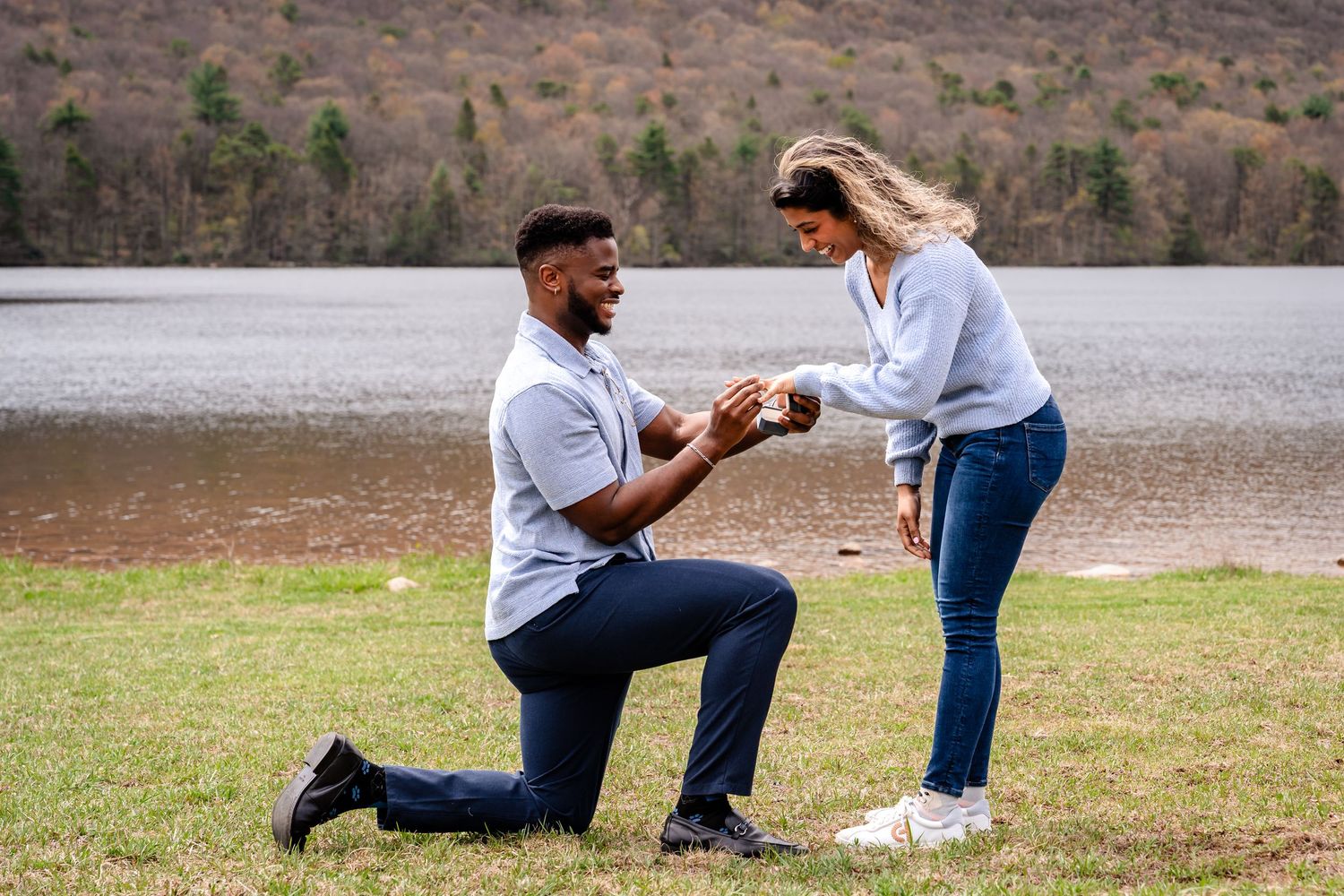 Man kneeling on one knee proposing to woman outdoors by a lake with scenic mountain backdrop.