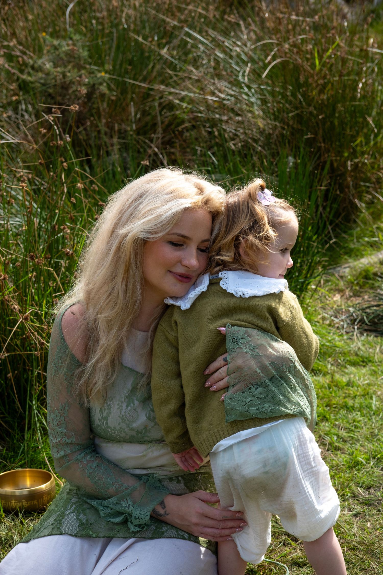 Blonde woman in green lace dress tenderly embracing a toddler outdoors in a sunlit grassy field.