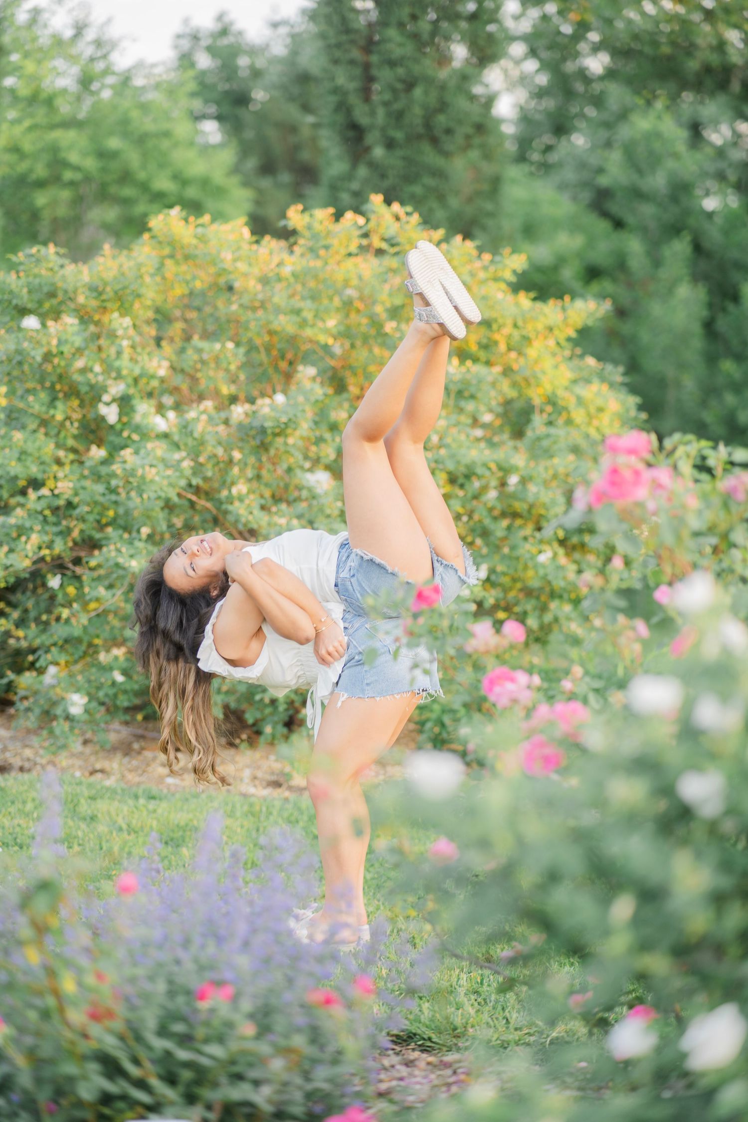 Young woman performing a high kick dance move in a colorful rose garden outdoors.