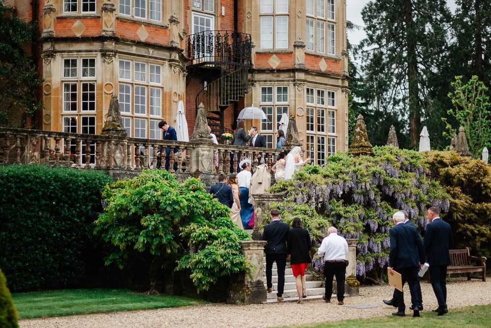 Elegant wedding guests gather outside a grand English manor adorned with wisteria blooms on a cloudy day.