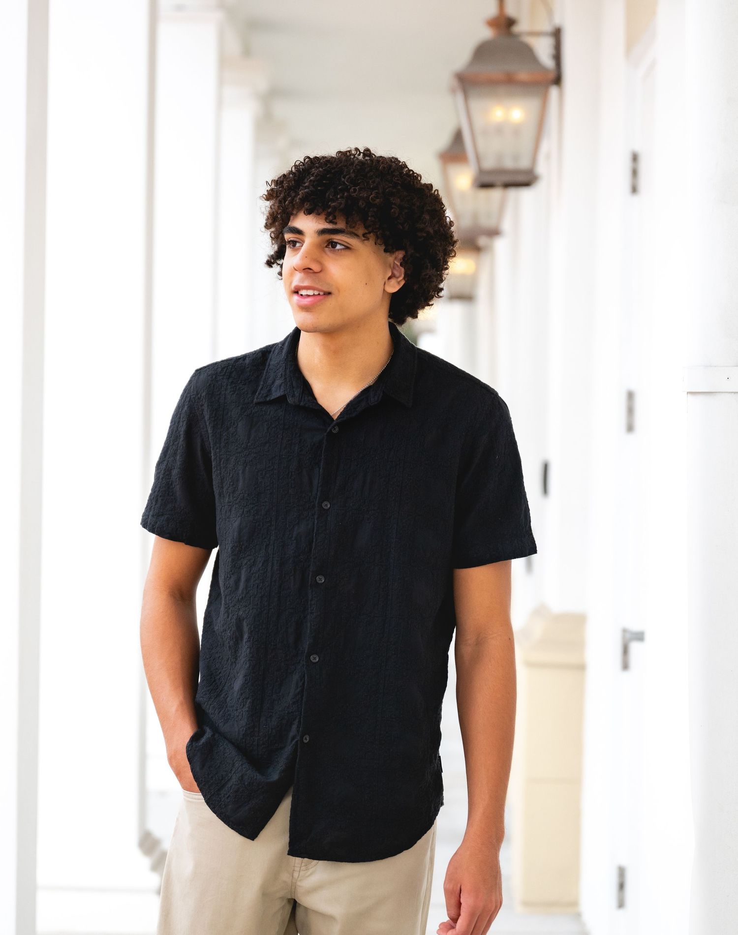 Young man wearing a black short-sleeve button-up shirt and khaki pants standing in a bright hallway.