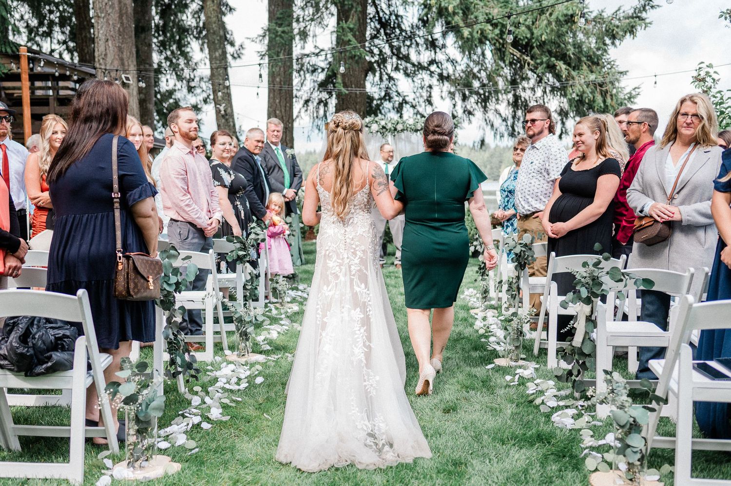 Bride walks down outdoor wedding aisle in lace gown, escorted by guest, with seated attendees on both sides.
