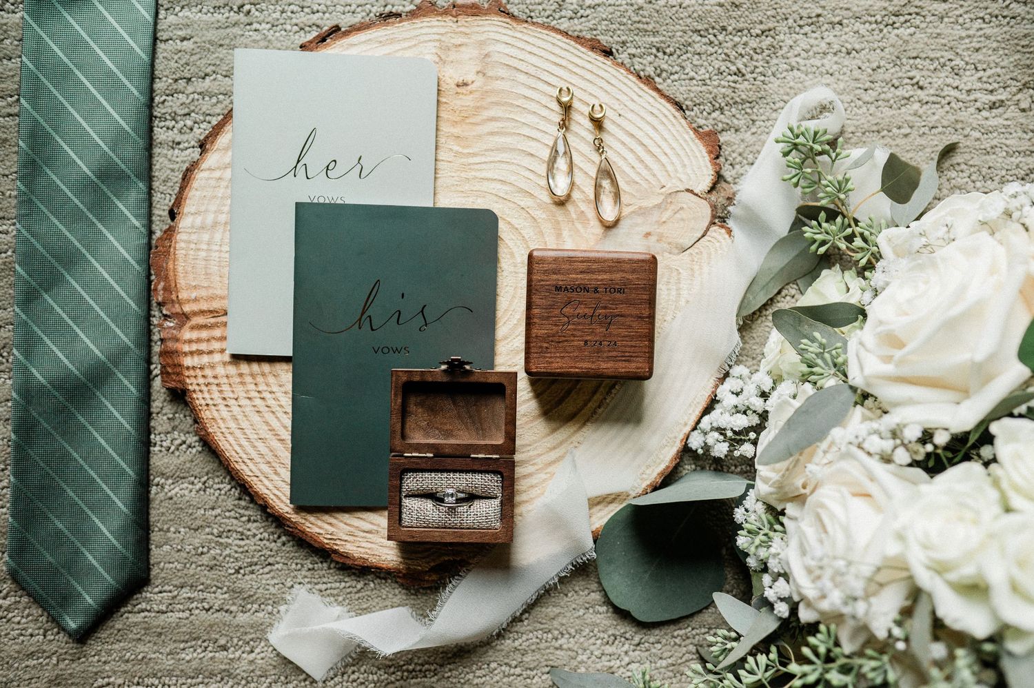 Wedding flat lay with green vow books, wooden ring box, diamond ring, earrings, green tie, and white roses on wood slice.