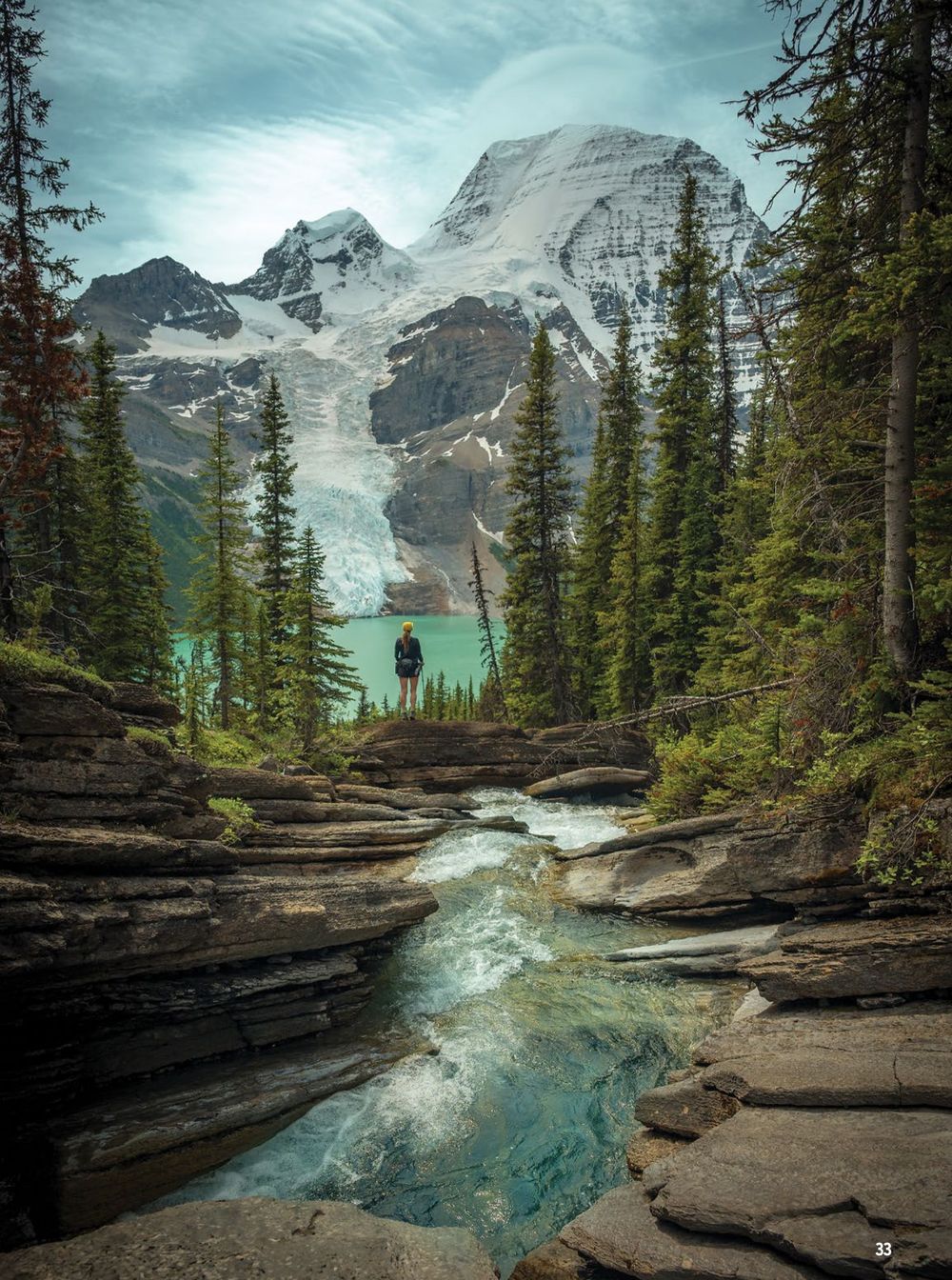 Rocky mountain trail winding through evergreen forest with turquoise alpine lake and snow-capped peaks.