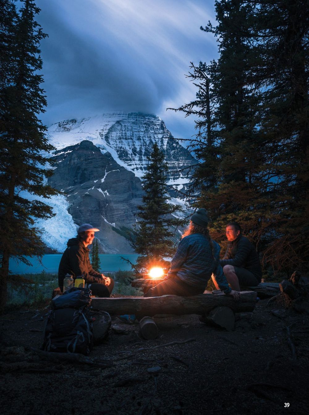 Campers gather around a glowing campfire at dusk with Mount Temple silhouetted against the moody blue sky in Banff.