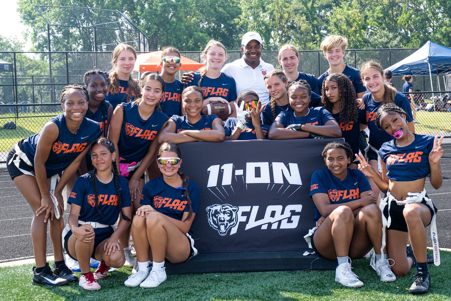 Girls flag football team poses with 11-ON FLAG banner on outdoor field during sunny tournament day.