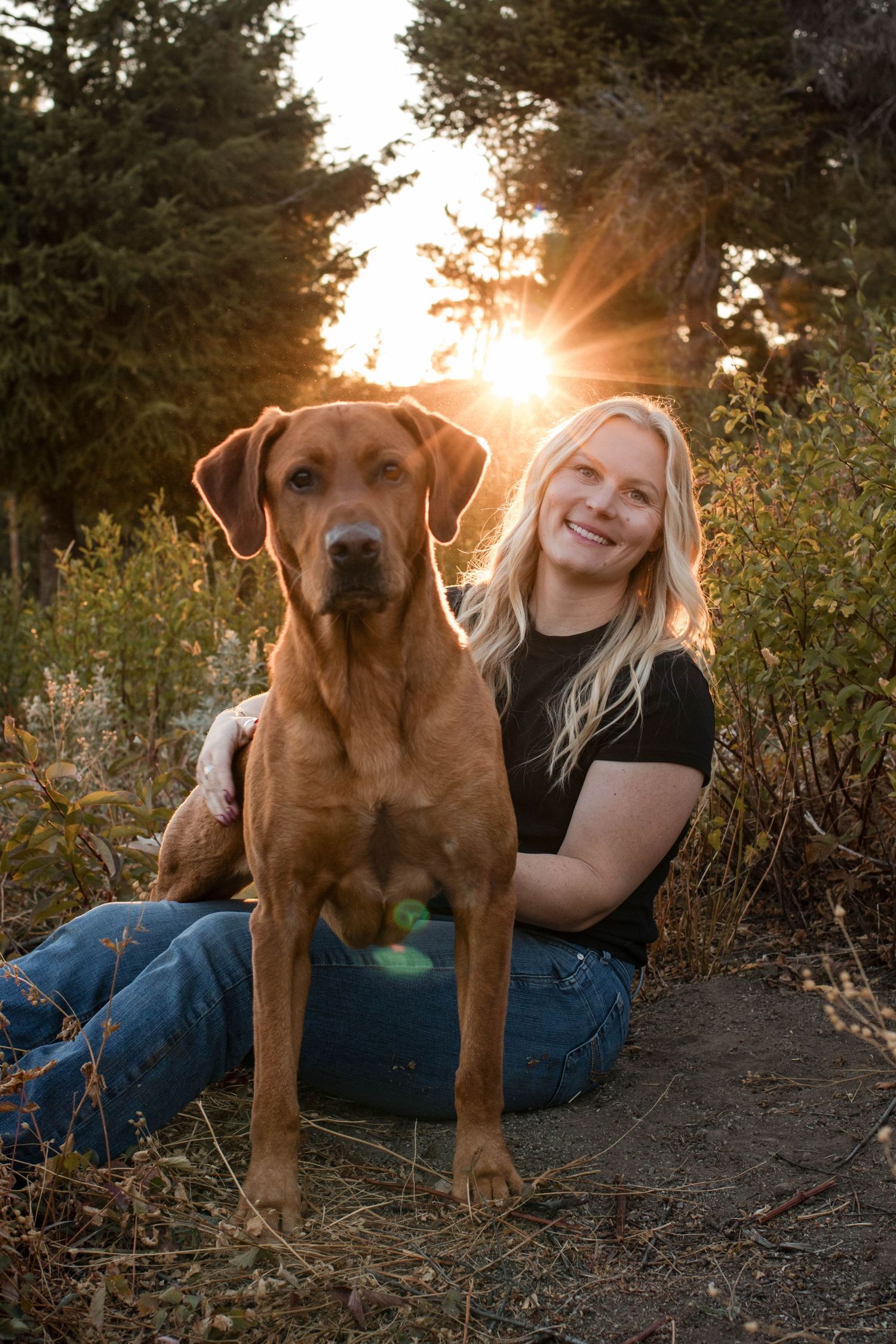 Smiling blonde woman sitting outdoors with a large brown Rhodesian Ridgeback dog at golden sunset.
