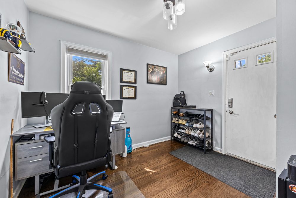 Home office with gaming chair, dual monitors, hardwood floors, and natural window light.