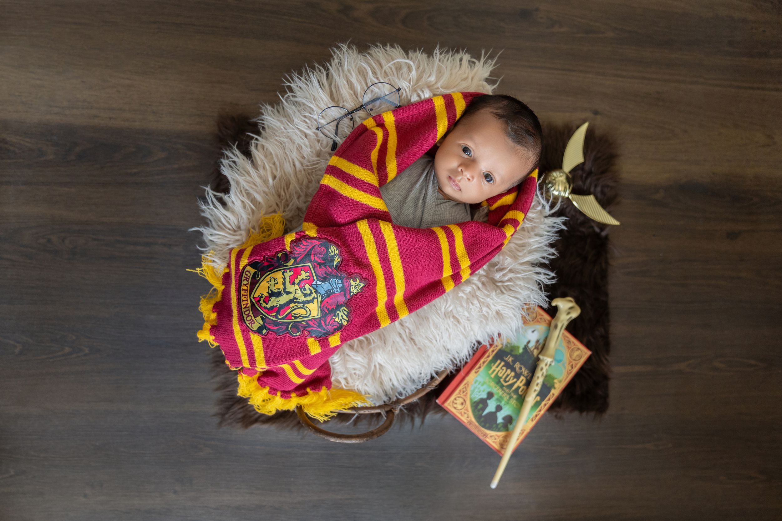 Newborn baby wrapped in a Gryffindor Harry Potter swaddle blanket, nestled in a fur-lined basket with a wand prop.