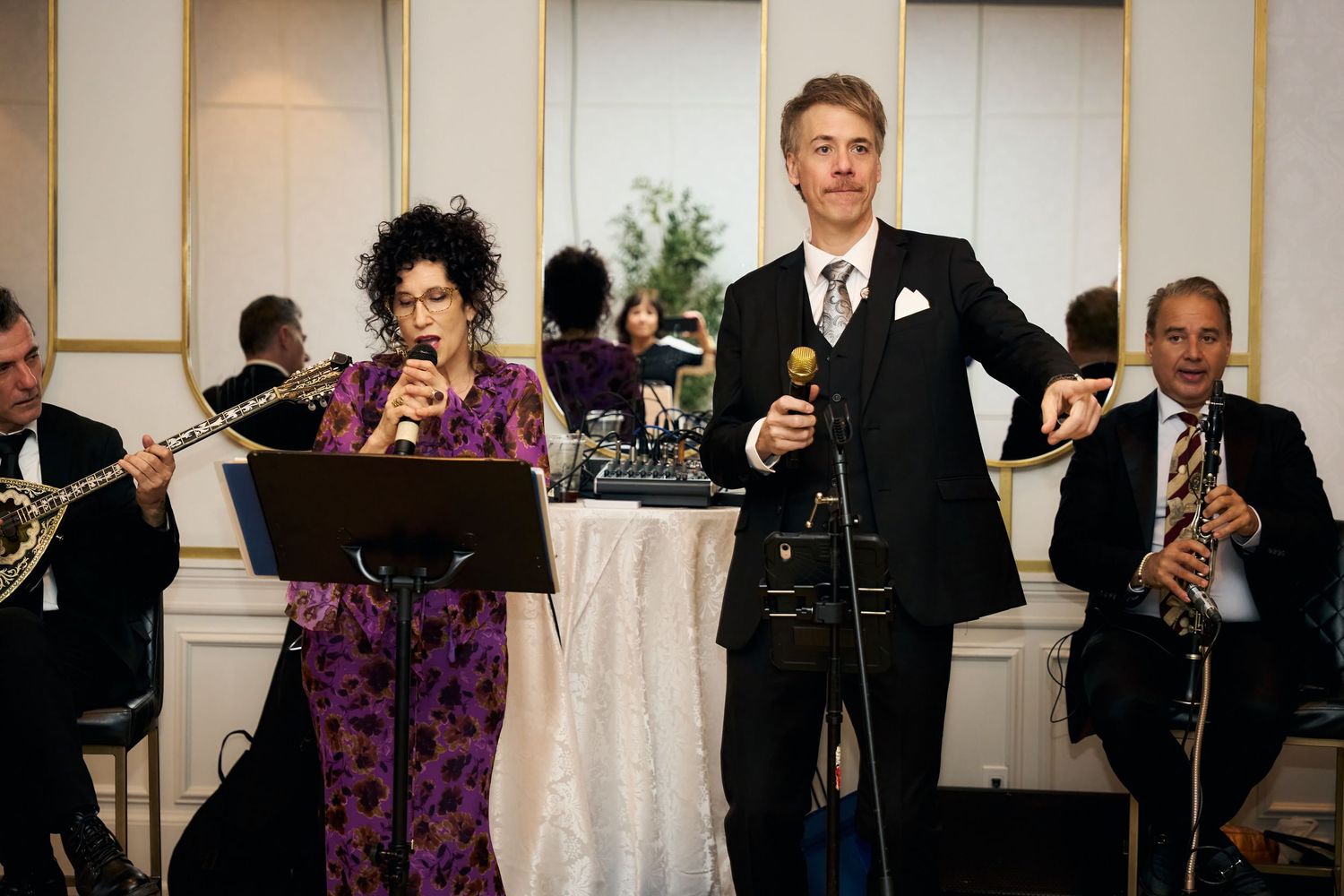 Man in black suit singing into microphone at formal event with saxophonist performing in background.