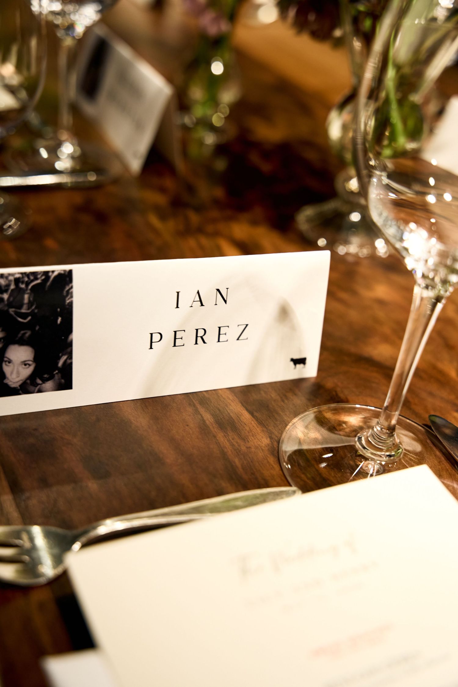 Elegant wedding place card for Ian Perez on a wooden table with wine glasses and cutlery at a formal dinner.