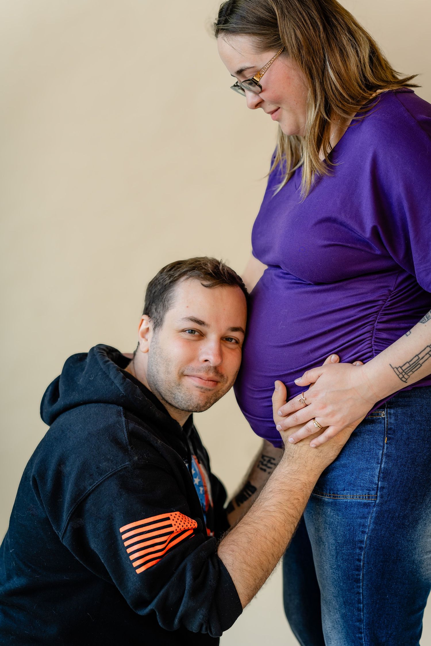 A man kneels beside a pregnant woman in a purple top, resting his head on her baby bump during a maternity photo session.