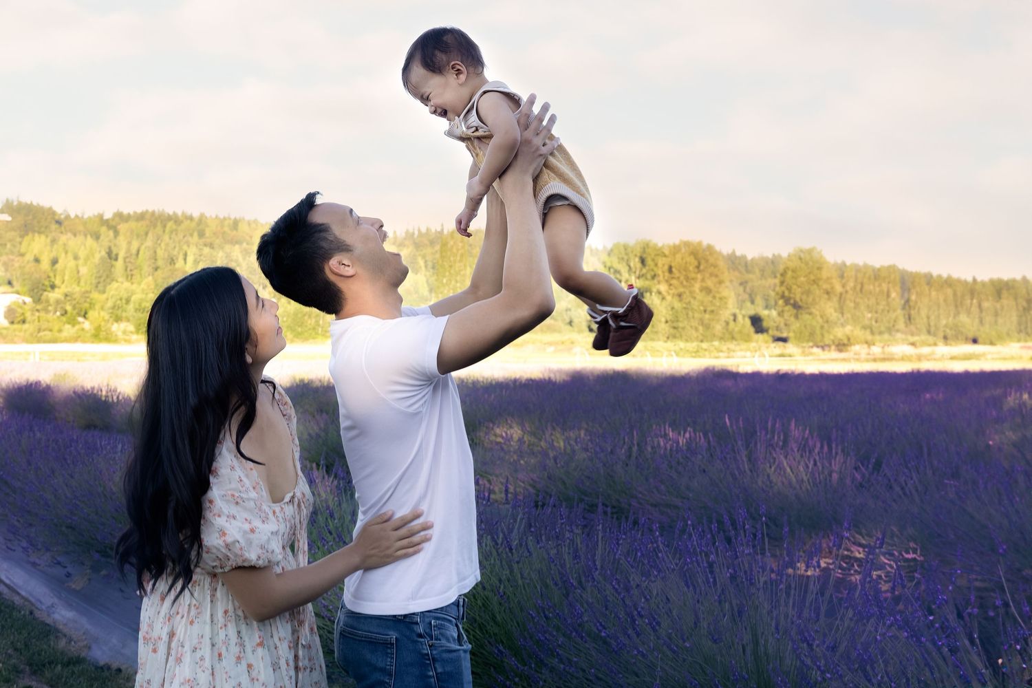A joyful family poses in a lavender field, with a man lifting a baby while a woman in a floral dress looks on.