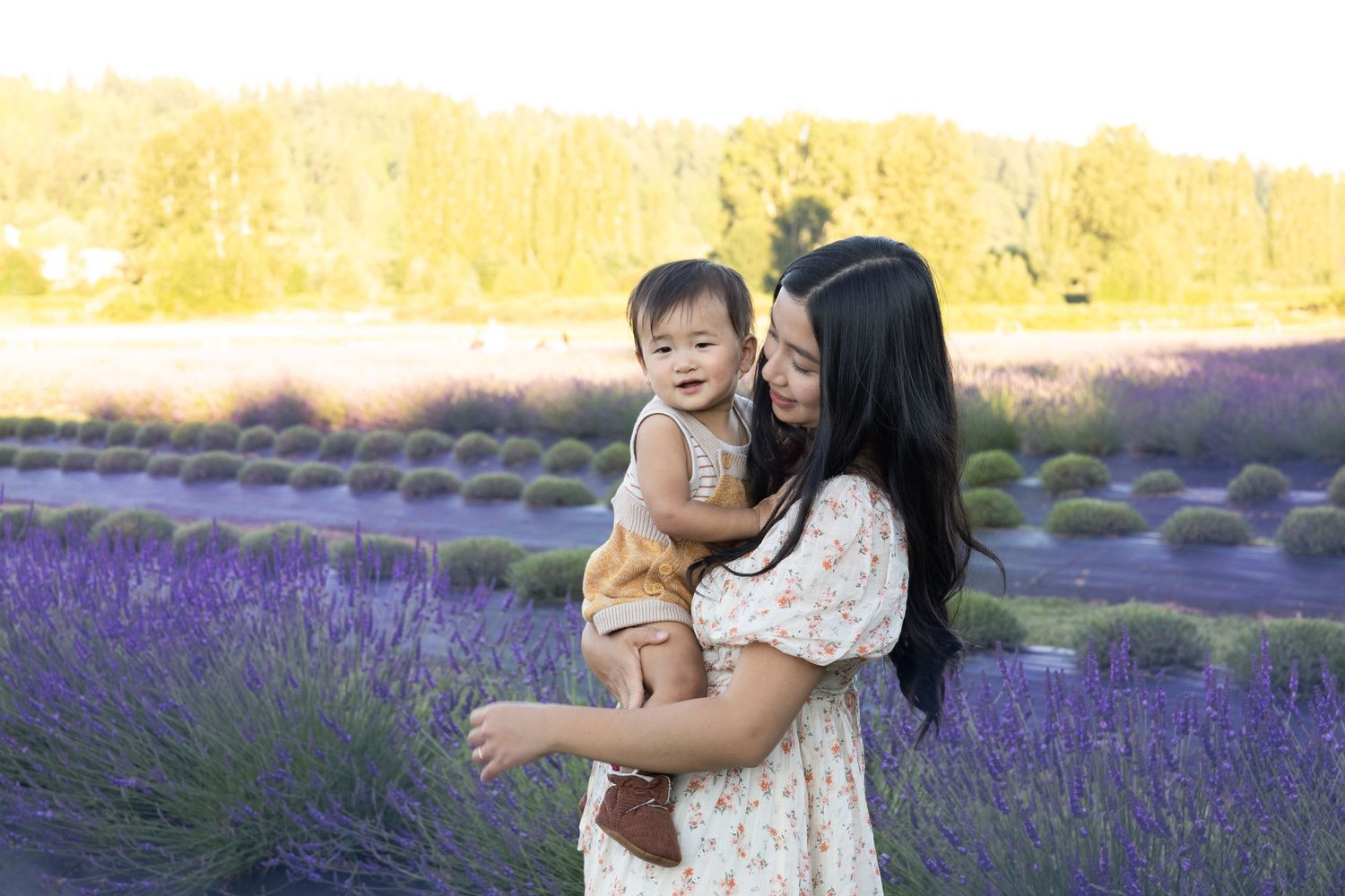 A mother holds her smiling toddler in a blooming lavender field at golden hour.