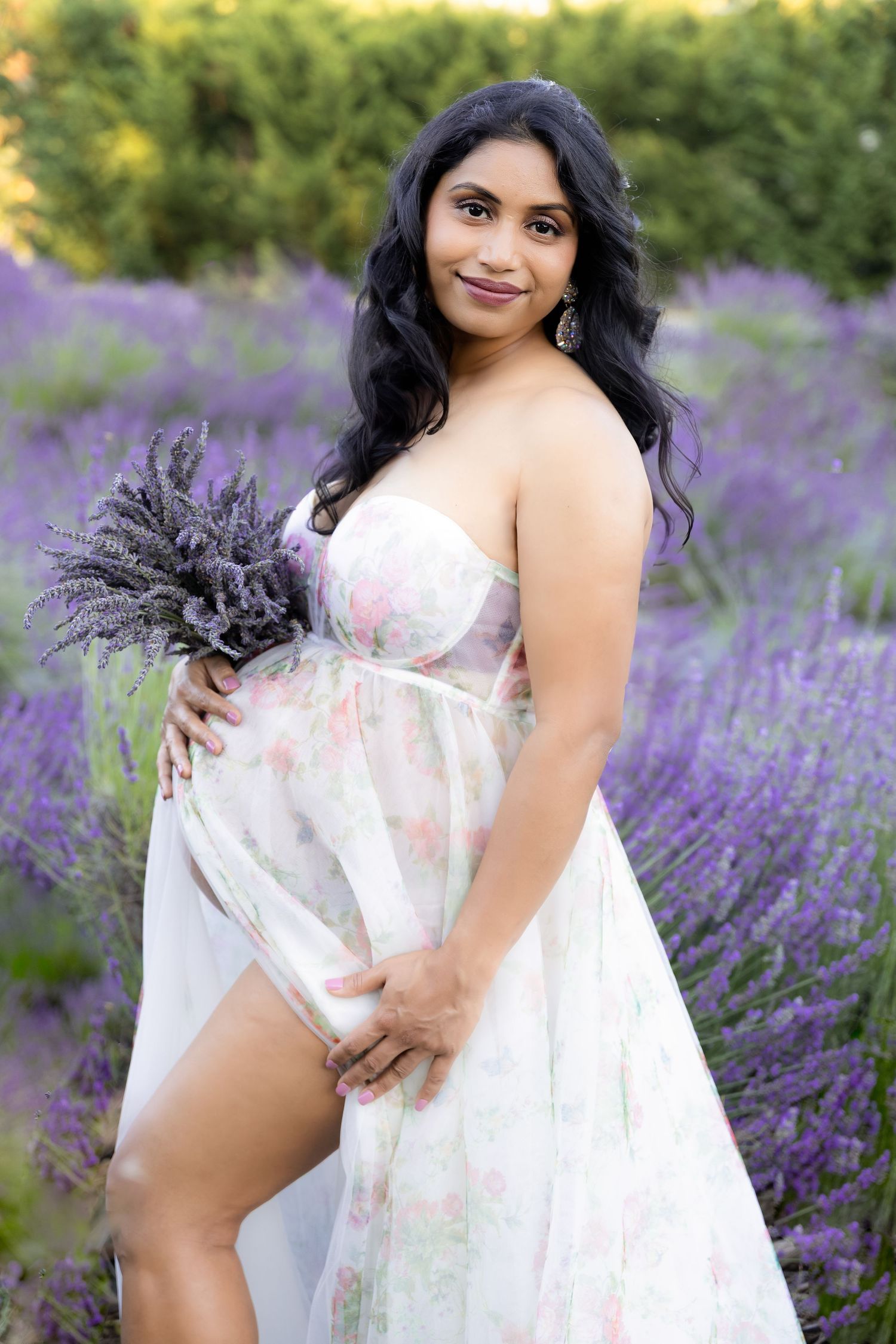 Pregnant woman in floral dress holding lavender bouquet, posing in a blooming purple lavender field.
