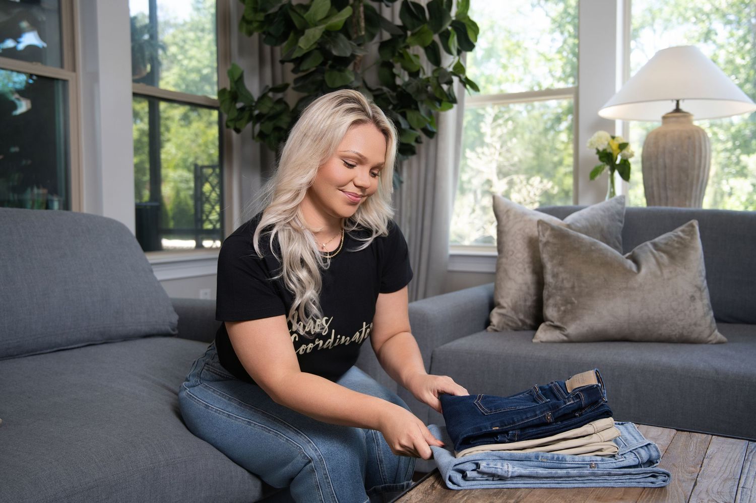Woman folding jeans on a coffee table in a cozy living room with a gray sofa and green plants.