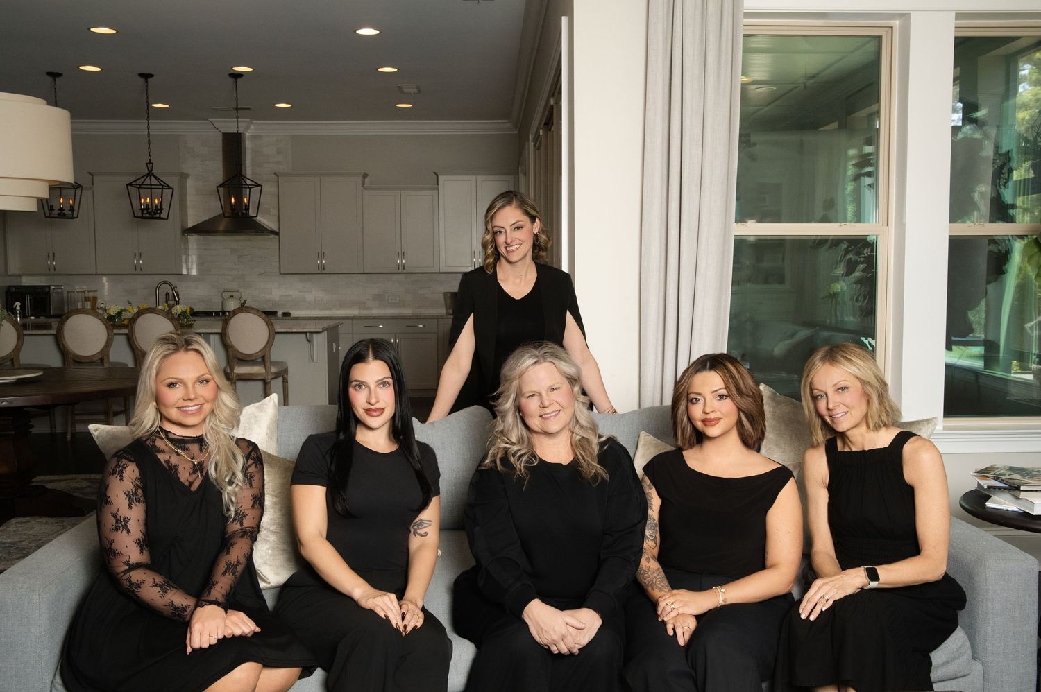 Six women in black outfits pose together in an elegant living room, five seated on a sofa and one standing behind.