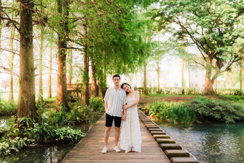 Couple posing on wooden boardwalk surrounded by lush green trees and serene water in a beautiful garden setting.