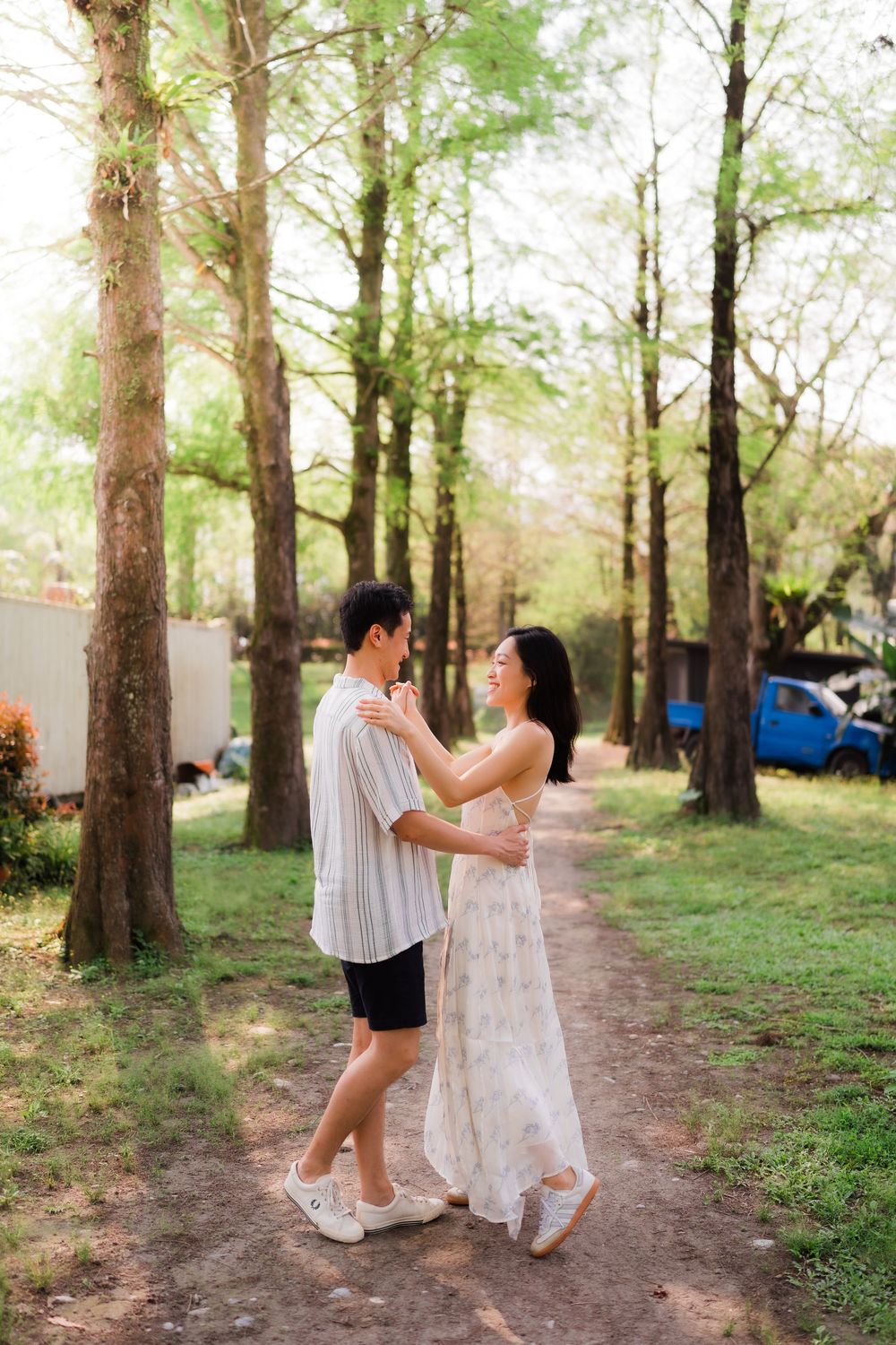 A couple dances together on a sunlit tree-lined path, the woman in a floral dress smiling at her partner.