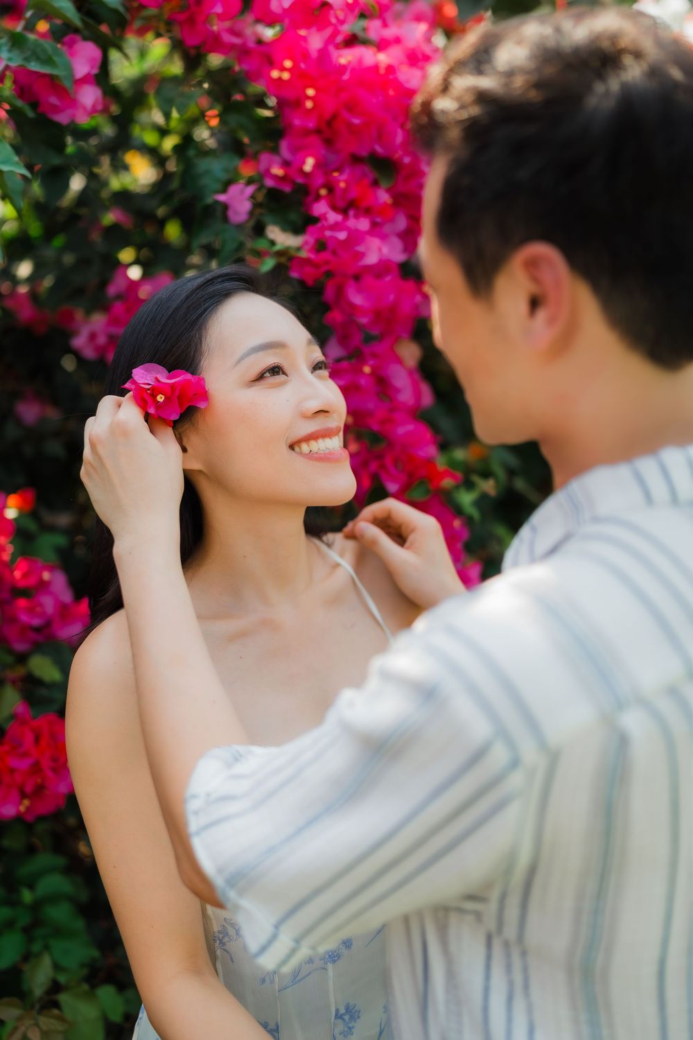A man places a pink flower in a smiling Asian woman's hair near vibrant bougainvillea blooms.