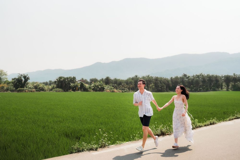 Couple holding hands running along a path beside lush green rice fields with mountains in the background.