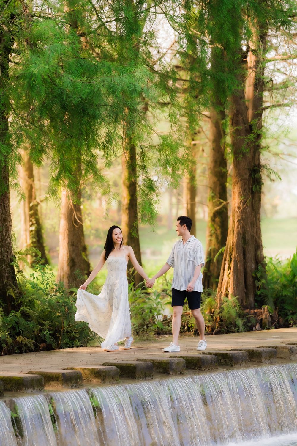 Couple holding hands walking on stone waterfall edge surrounded by tall green trees in a lush forest setting.