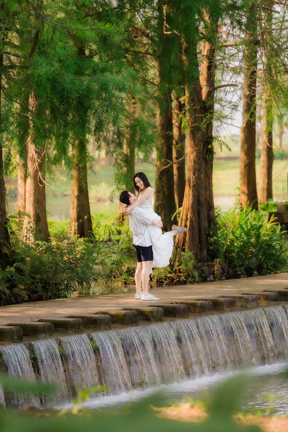 A man lifts a woman in a white dress on a wooden bridge over a waterfall in a lush forest.
