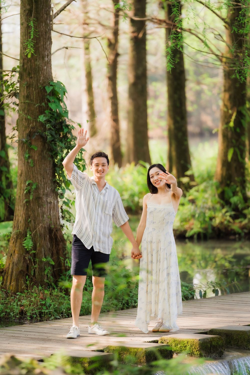 Happy couple holding hands and waving on a wooden bridge surrounded by tall trees and a serene pond.