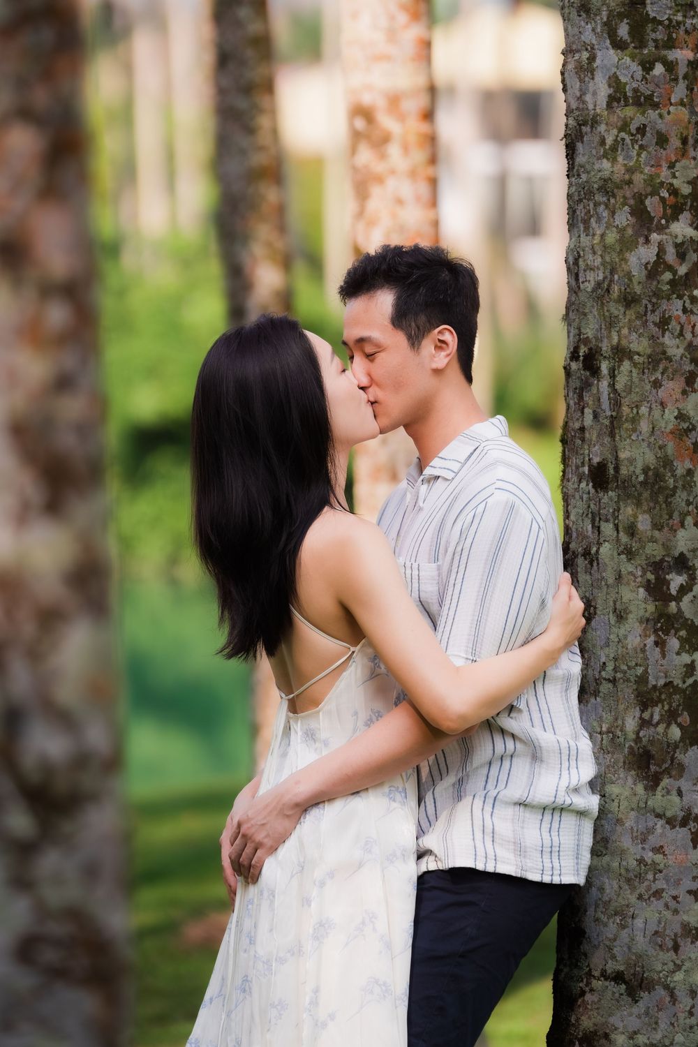 An Asian couple shares a tender kiss leaning against a tree trunk in a green park setting.