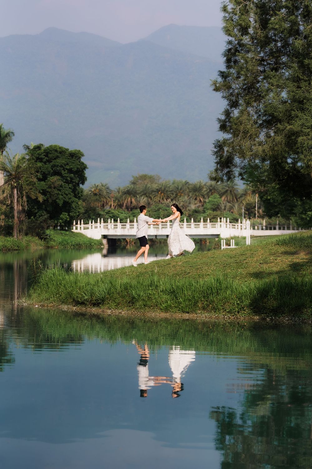 Couple dancing on white bridge reflected in calm lake with mountains and palm trees in background.