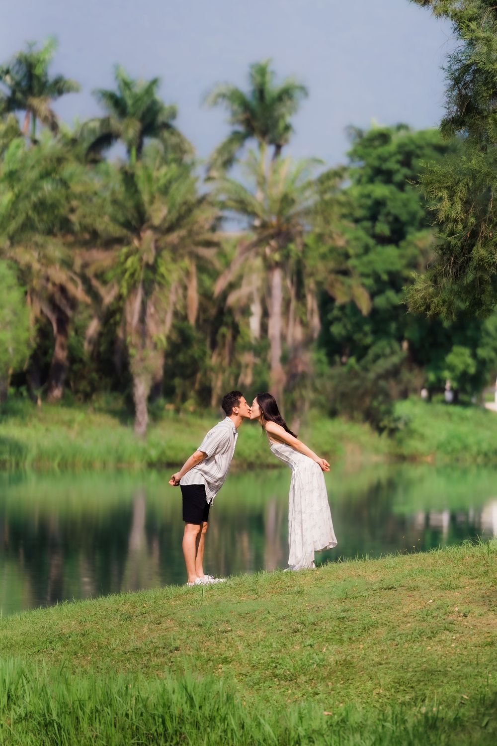 Asian couple sharing a romantic kiss by a serene pond surrounded by lush tropical greenery.