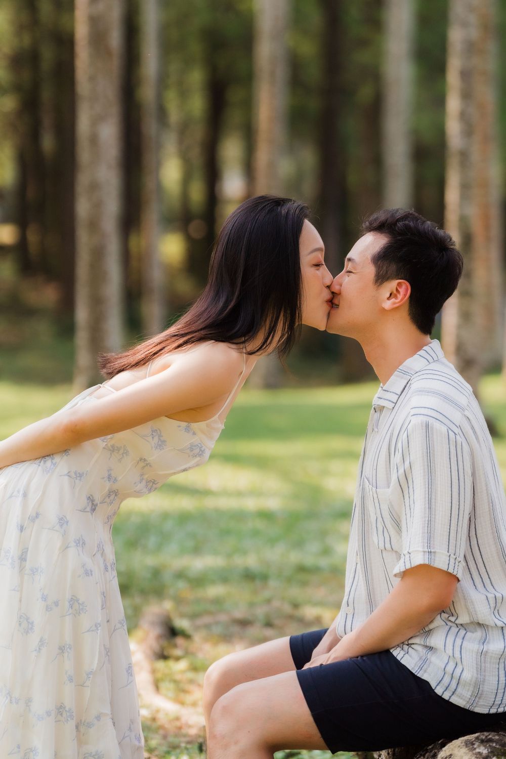 Asian couple sharing a tender kiss in a sunlit forest park setting, woman in floral dress.