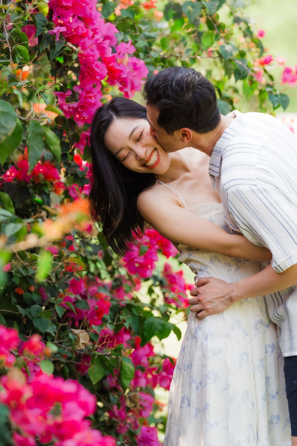 A man kisses a laughing woman on the cheek while embracing her near bright pink bougainvillea flowers.