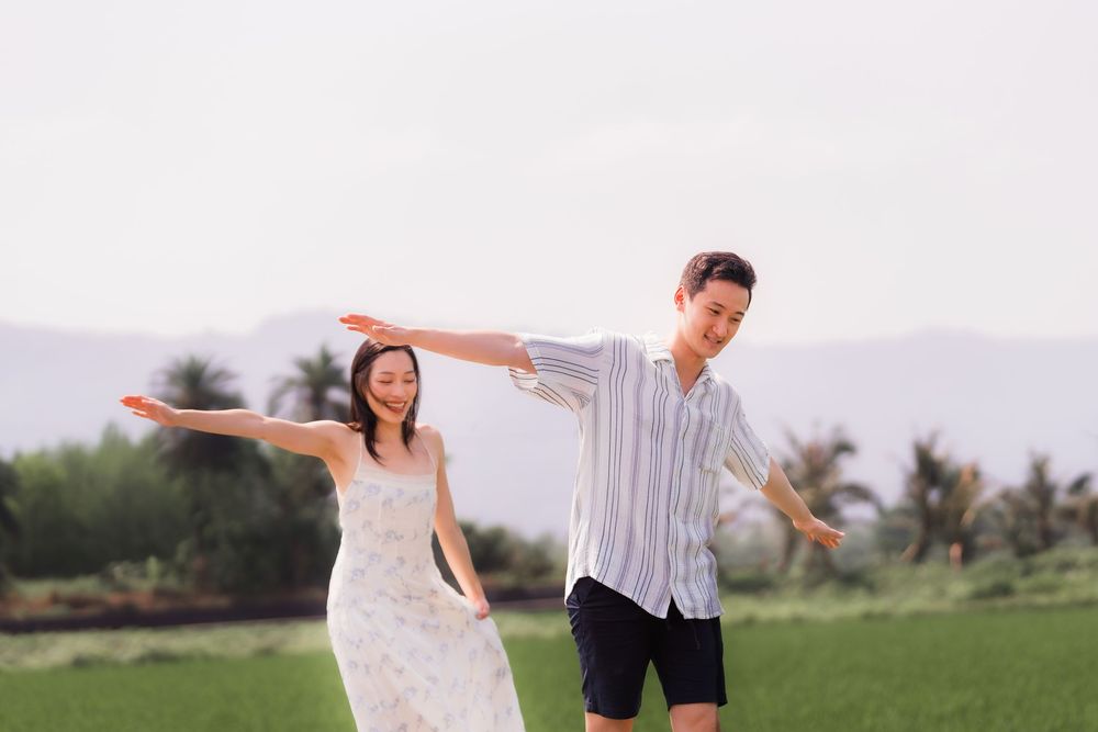 A joyful couple spreads their arms wide in a lush green field with tropical trees in the background.