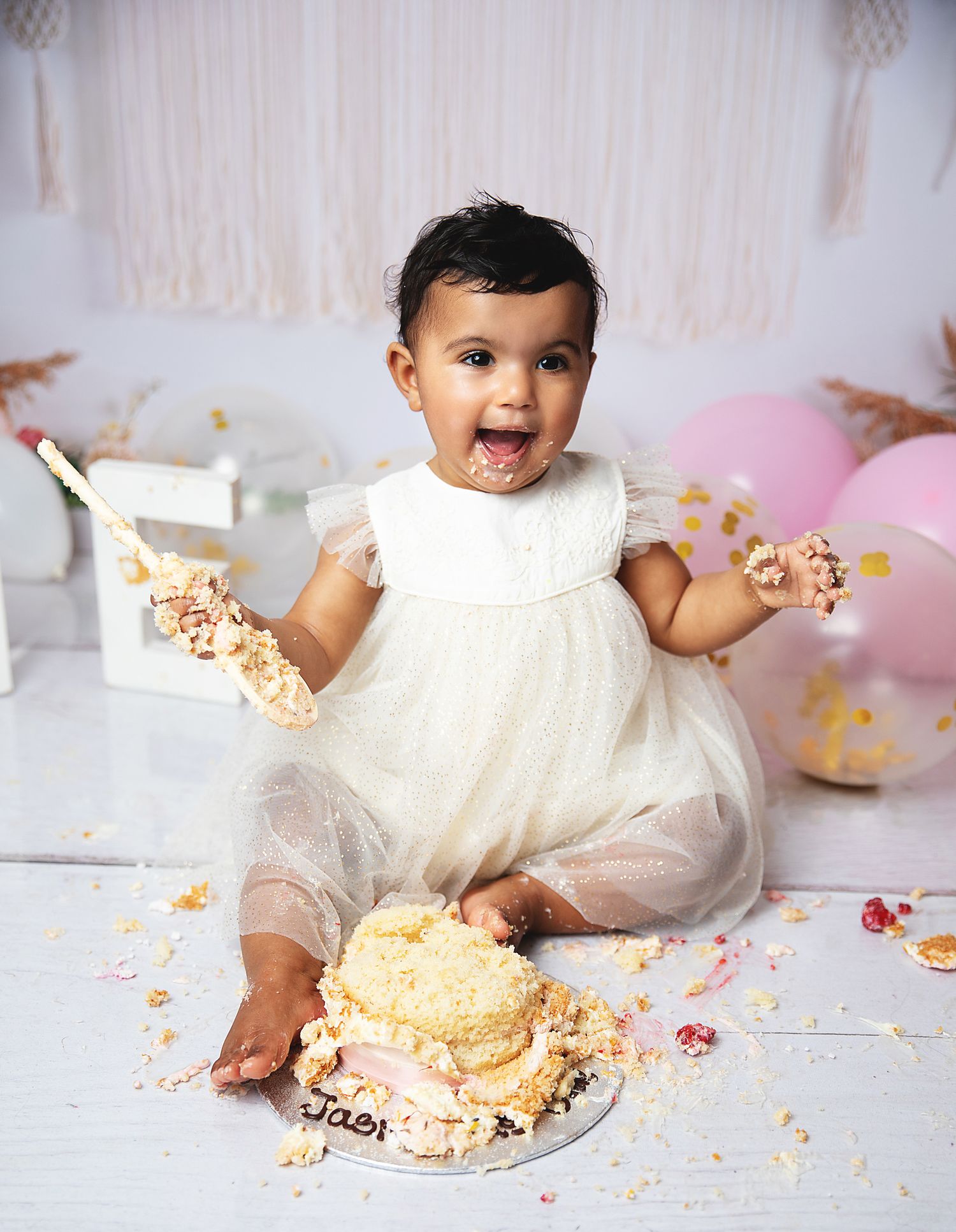 Happy baby in white dress messily enjoying birthday cake smash photo session with pink and gold balloons during a photoshoot in Bexley