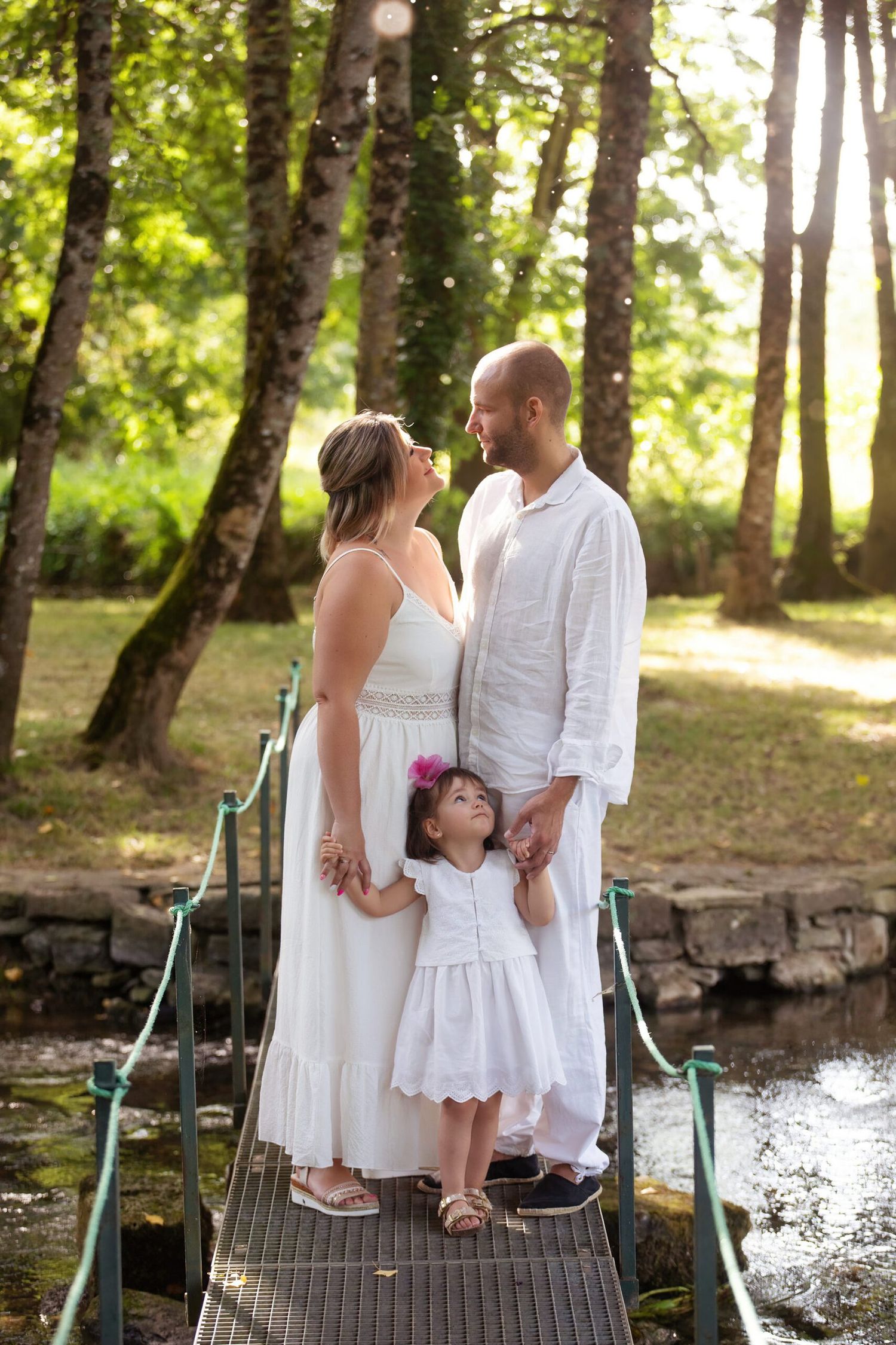 Family wearing white outfits stands together on a wooden dock in a sunlit forest setting near water.