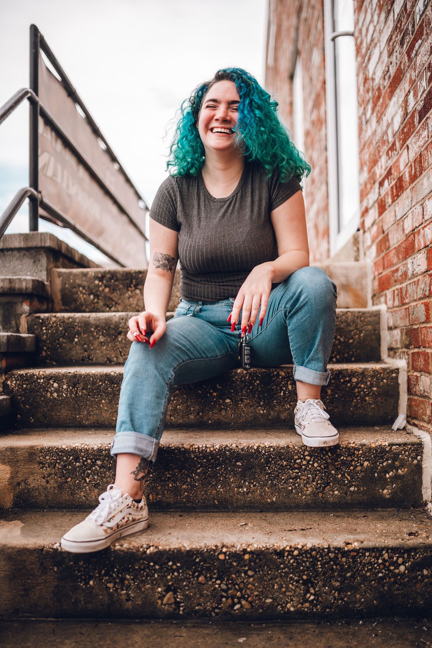 Person with vibrant turquoise hair sitting casually on concrete steps wearing jeans and sneakers while smiling outdoors.