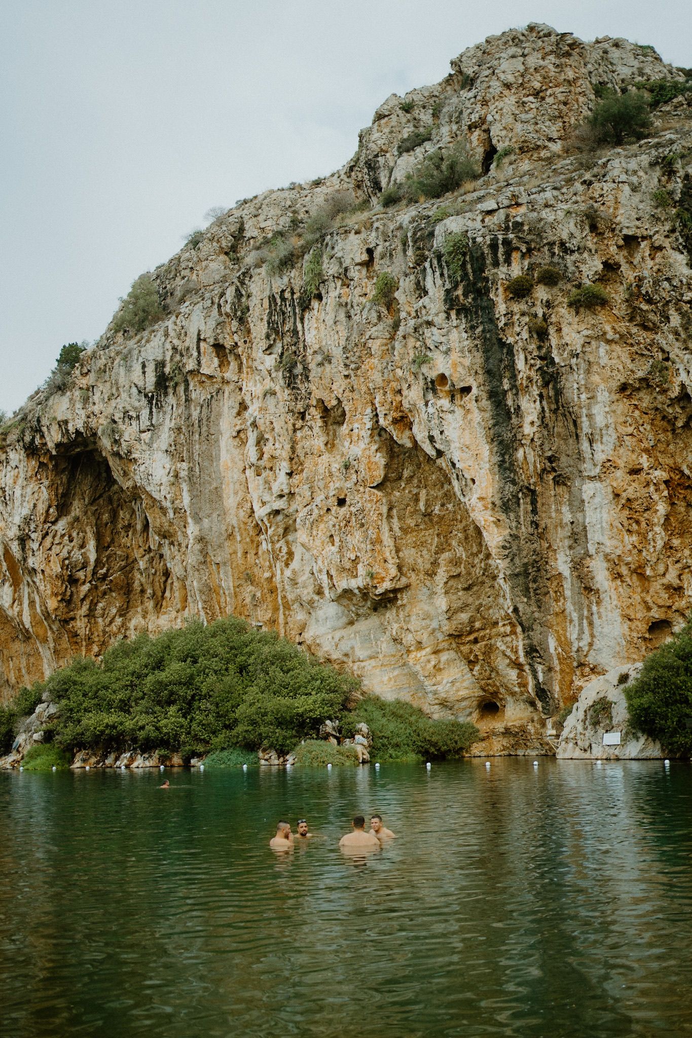 Steep limestone cliff face rises above tranquil green lake with swimmers enjoying the water on a cloudy day.