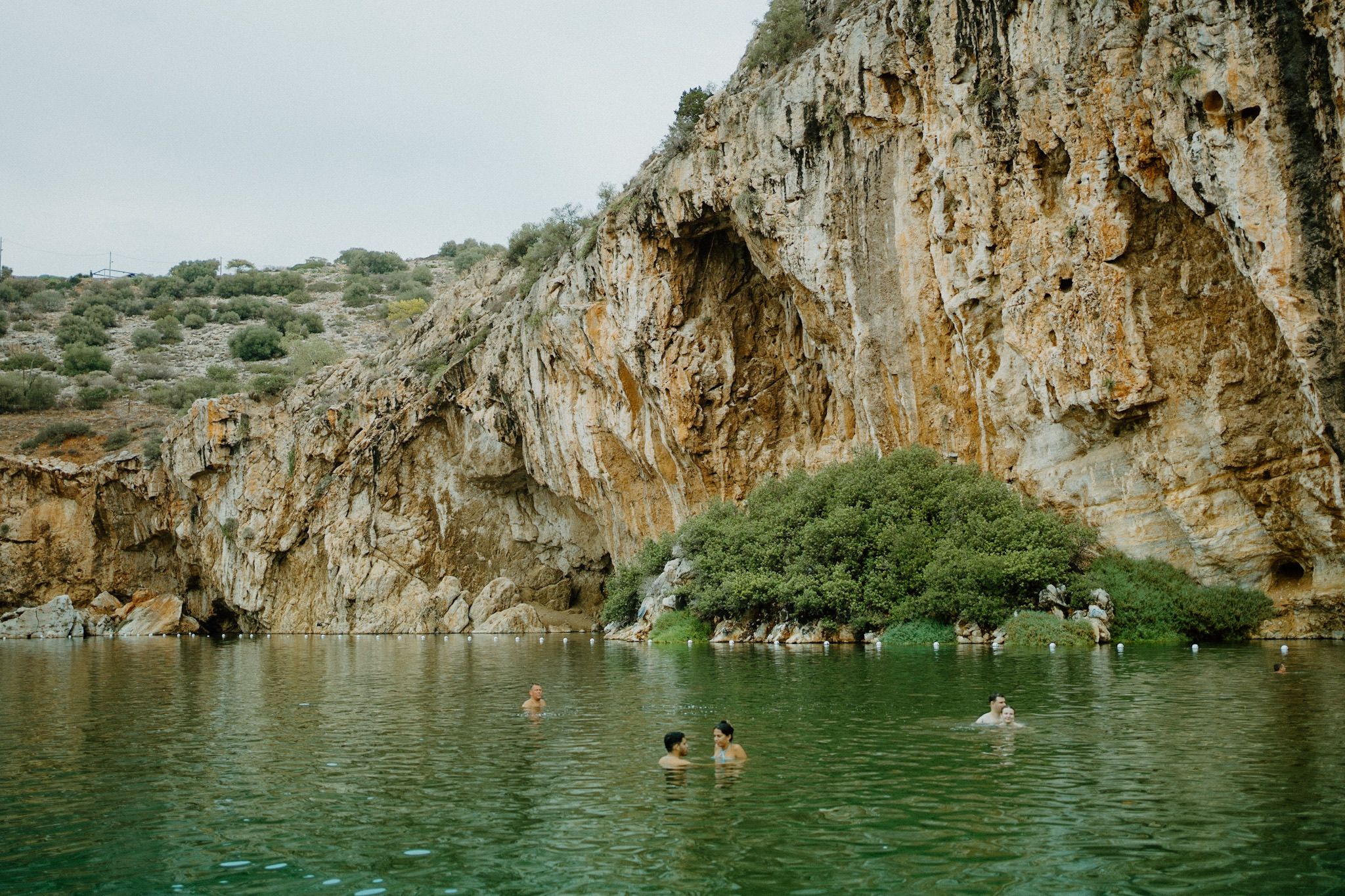 People swim in turquoise water beneath tall limestone cliffs at a natural swimming hole on a summer day.