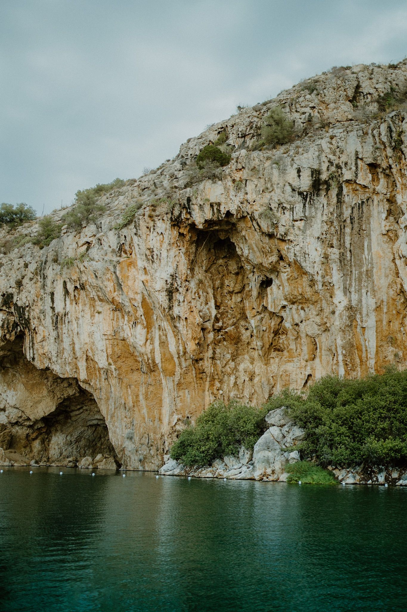 Steep limestone cliffs rise above dark teal waters at a Mediterranean lake with green vegetation near the base.