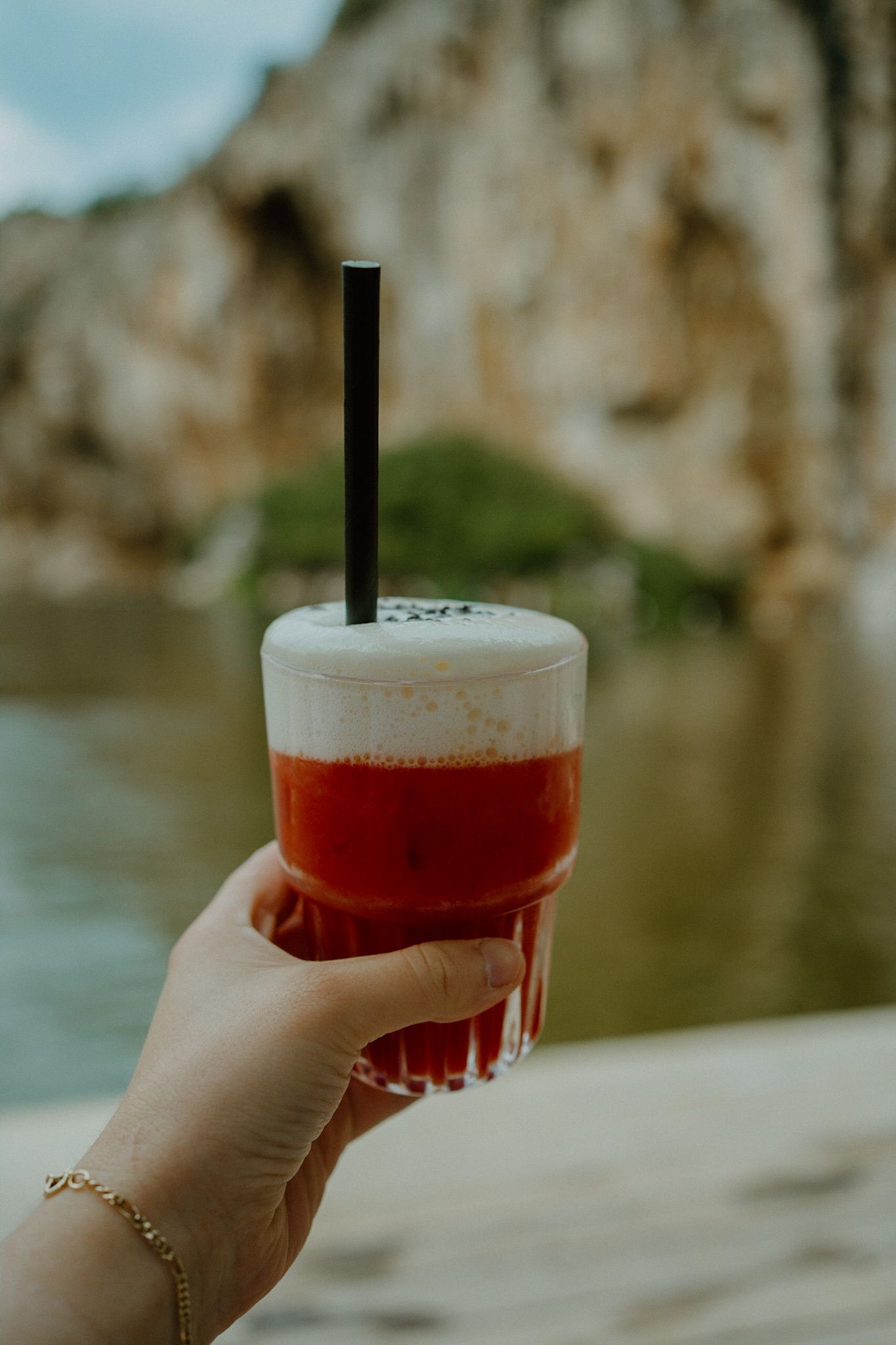 Hand holding a vibrant red layered drink with foam and black straw against a blurred rocky cliff and water background.