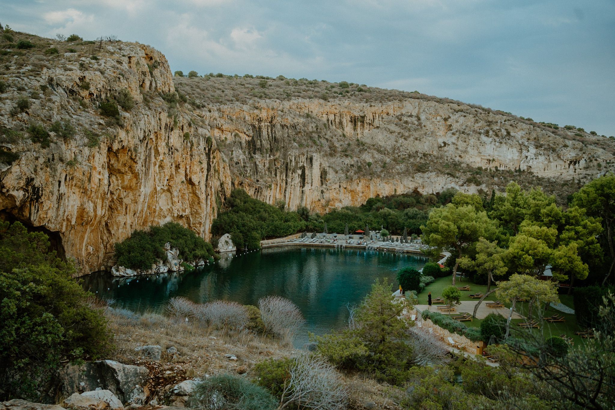 A scenic bridge spans a tranquil lake surrounded by dramatic limestone cliffs and lush greenery in Vouliagmeni, Greece.