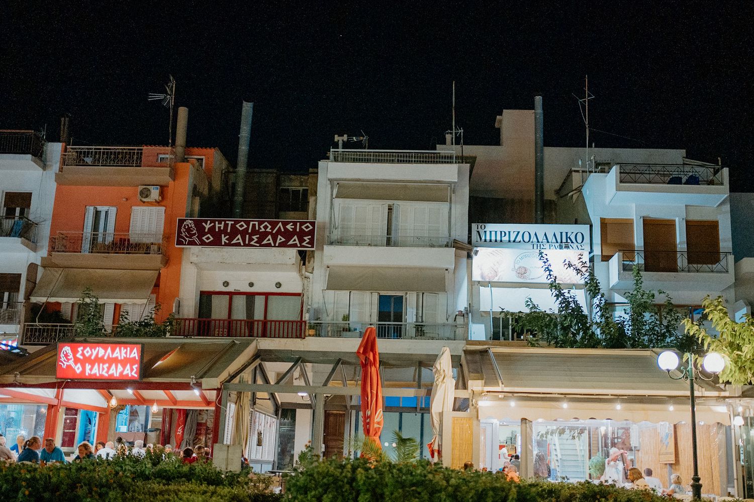 Night view of illuminated storefronts and buildings along a Mediterranean street with neon signs and outdoor seating.