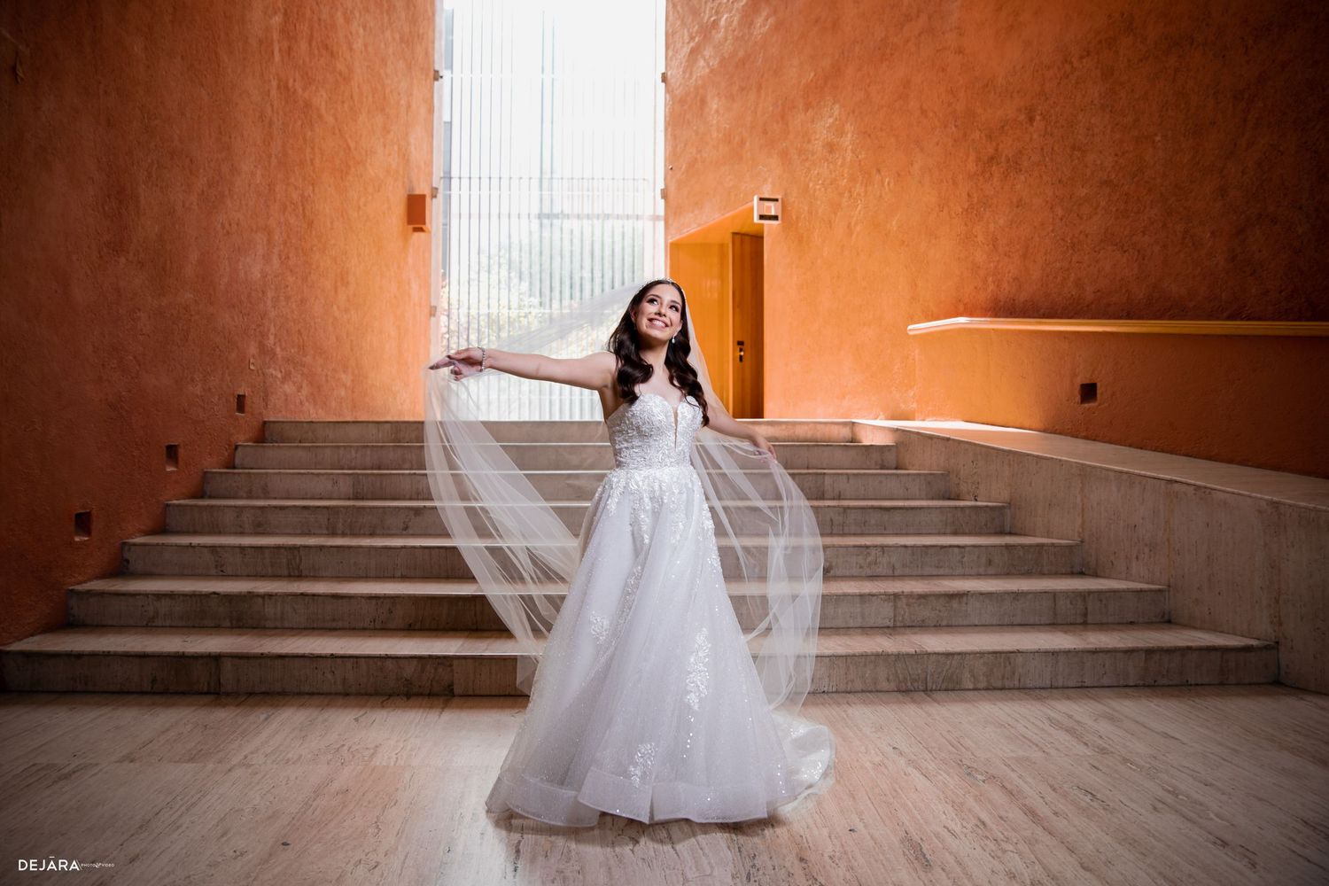 A bride in a white gown poses on grand stone stairs in a warm-toned hallway.