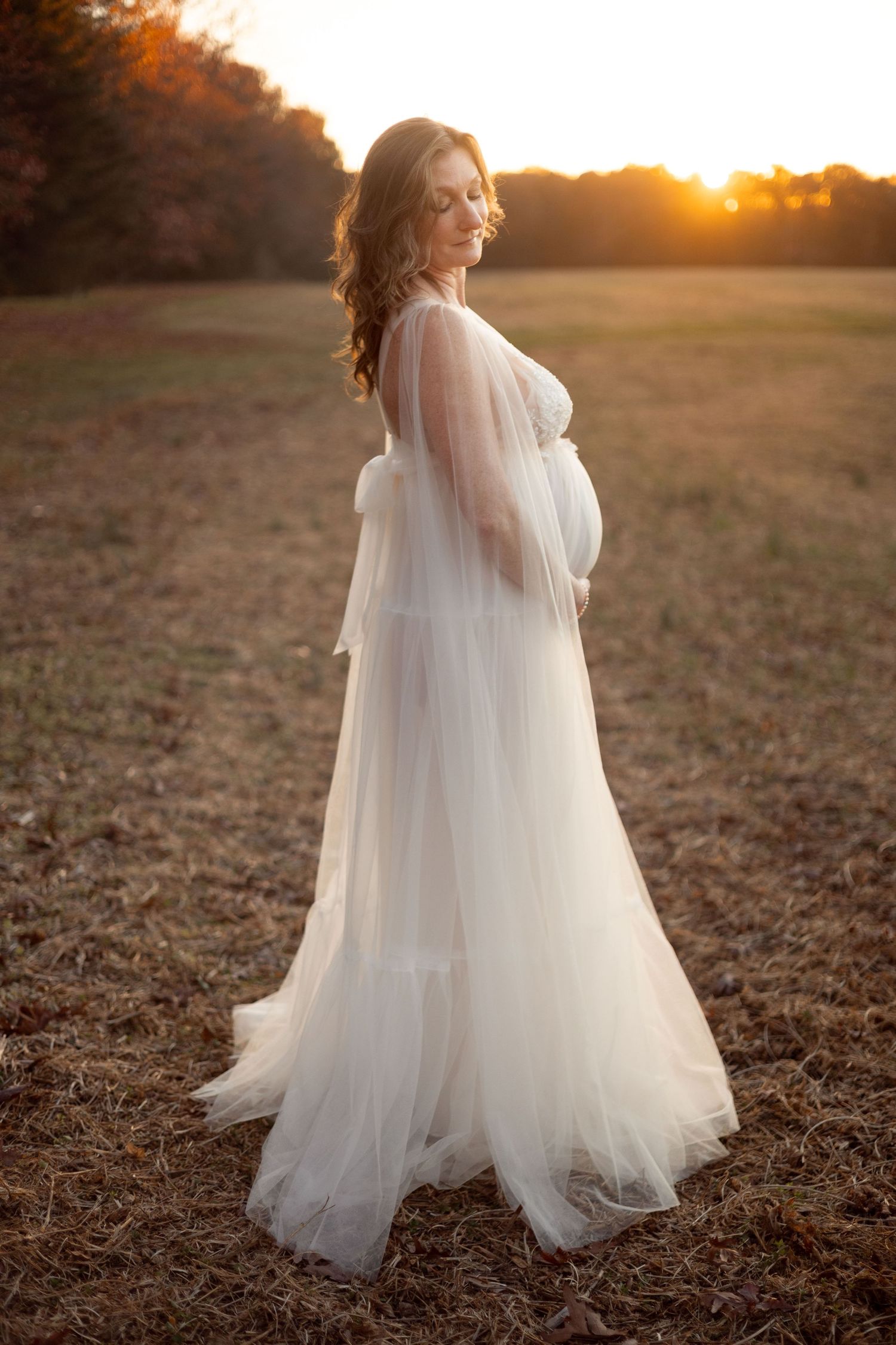 Pregnant woman stands in a field at sunset.