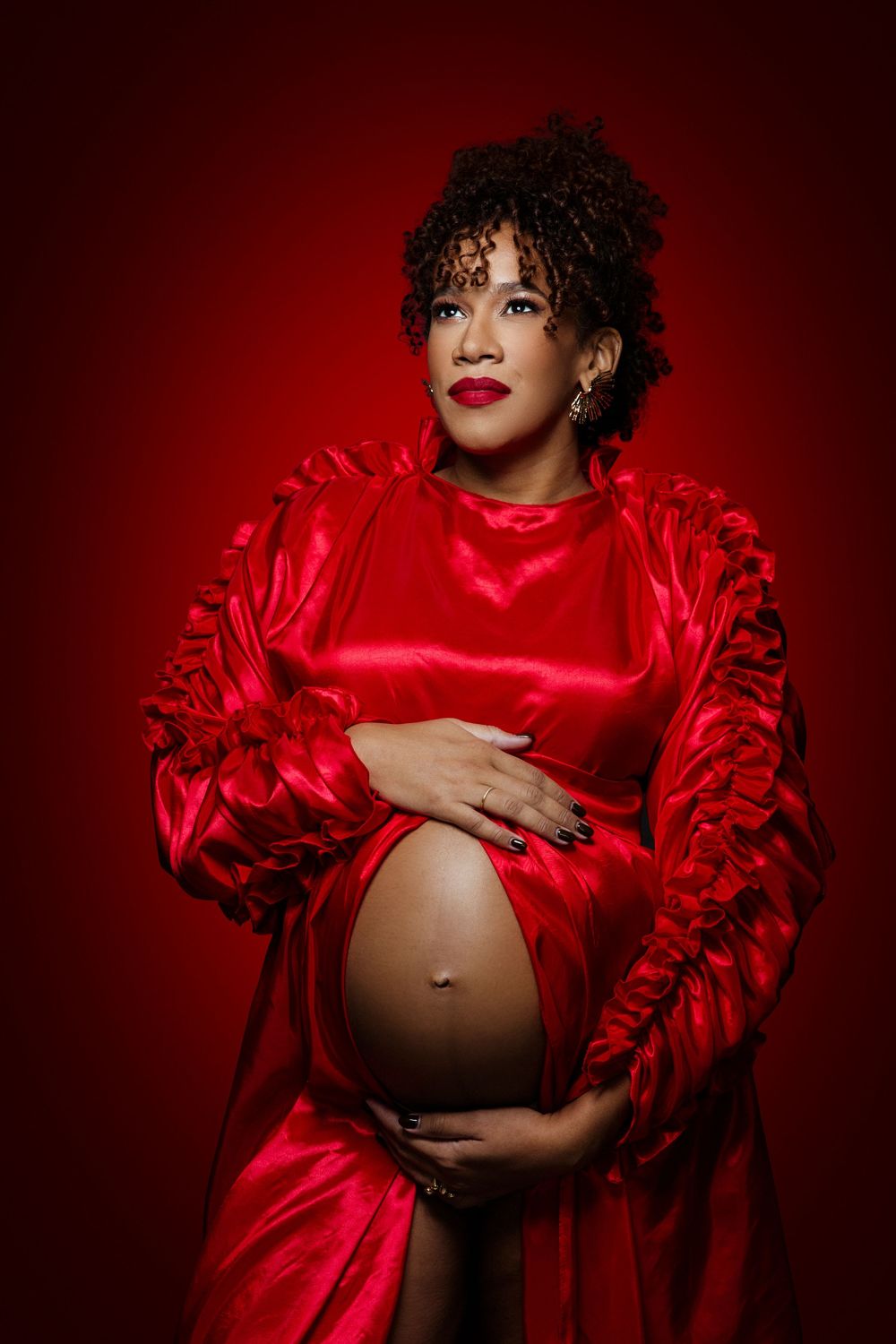 Pregnant woman in red satin robe cradling baby bump against red background, maternity portrait photography.
