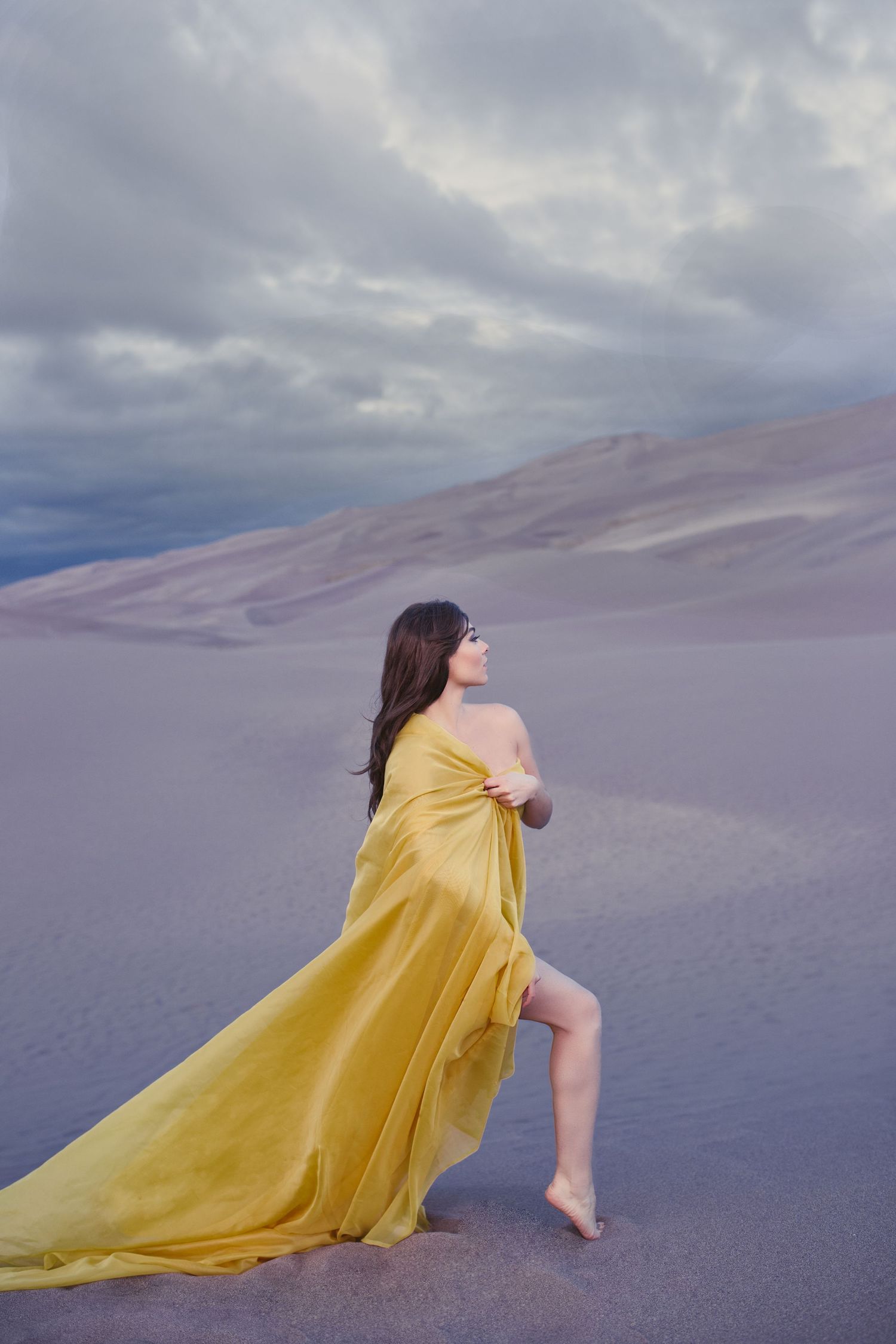 Figure in flowing yellow dress stands gracefully on sand dunes under moody cloudy sky at sunset.