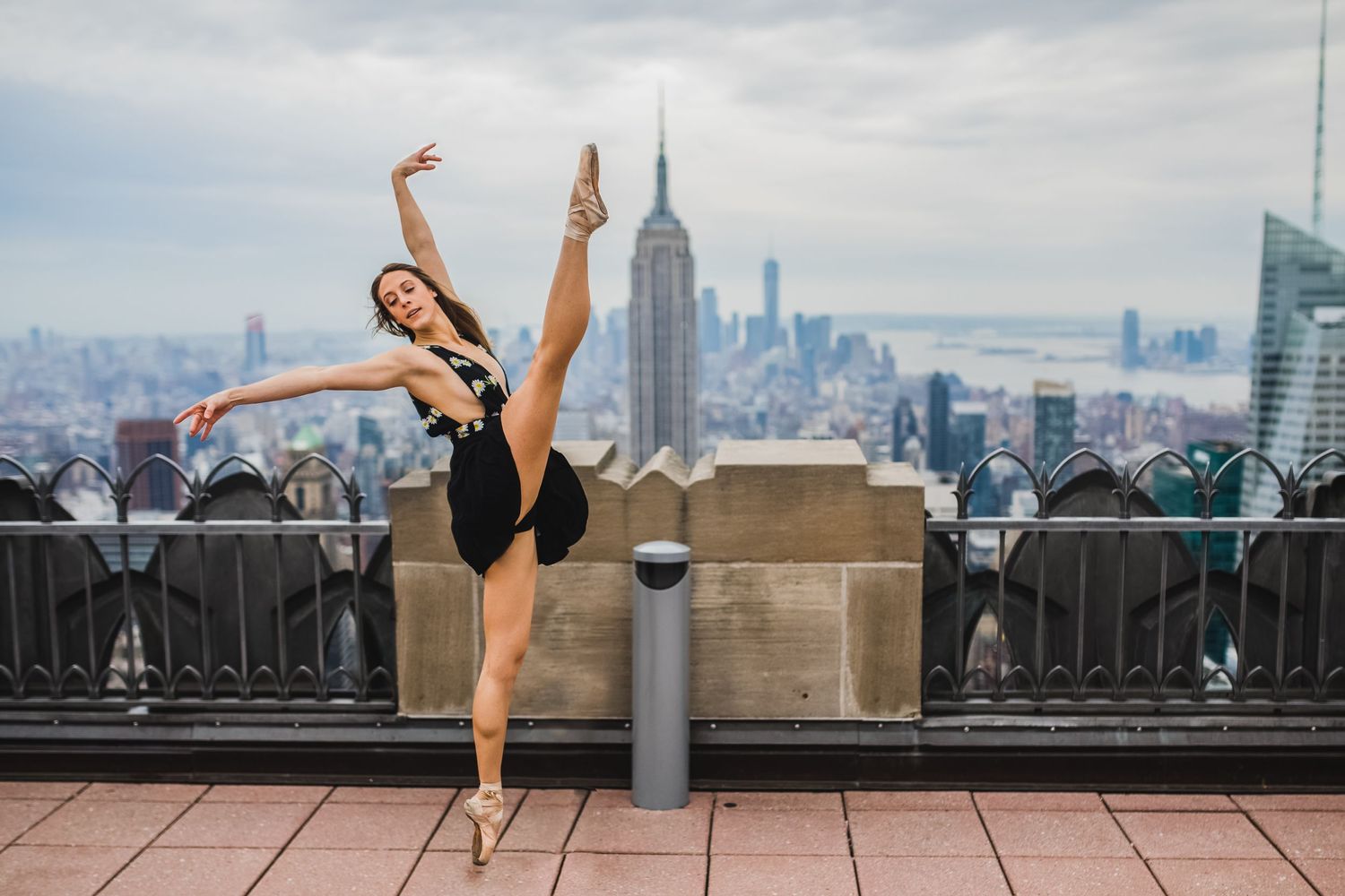 A dancer performs a graceful arabesque pose on an observation deck overlooking the Empire State Building and NYC skyline.