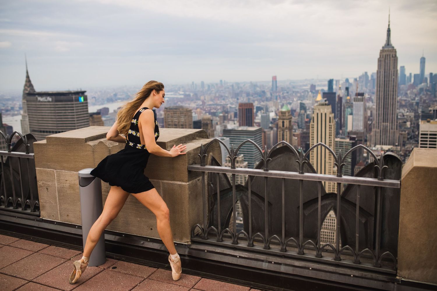 A dancer in a black dress performs an elegant pose atop a skyscraper observation deck overlooking the New York City skyline.
